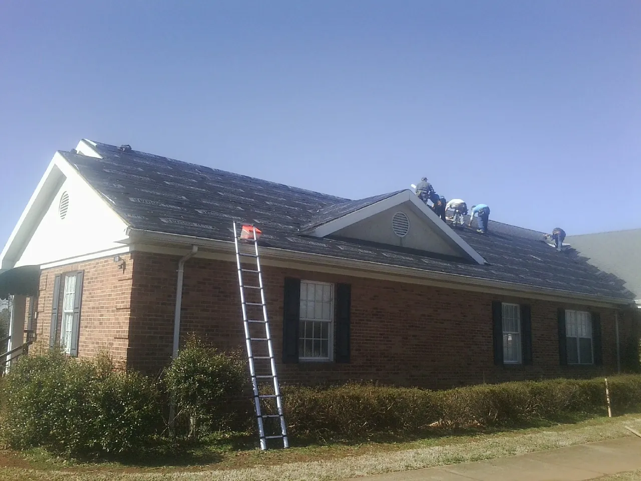 Crew tearing shingles off a brick ranch roof with a ladder set — Metal Roof Repair work by our crew in Goshen